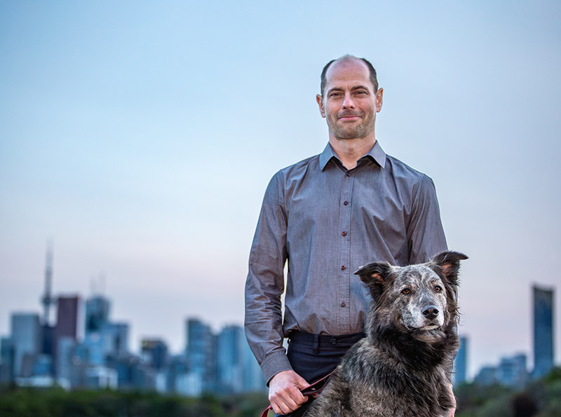 Dog sits in front of man with blurred imaged of Toronto skyline in background